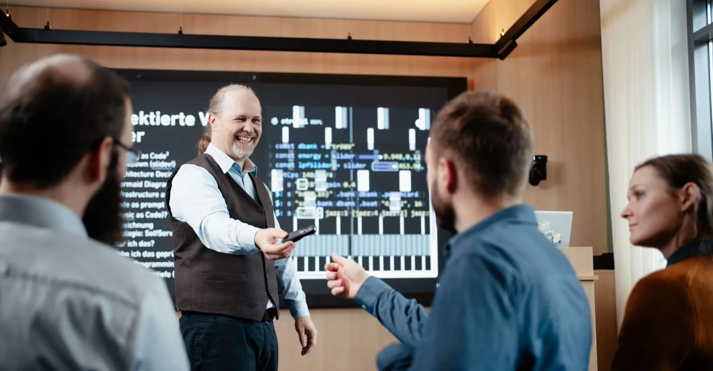 Ein Berater der viadee steht vor einem Smartboard und reicht lächelnd den Pointer einem Kollegen. Zwei weitere viadee Berater:innen sitzen links und rechts im Bild.