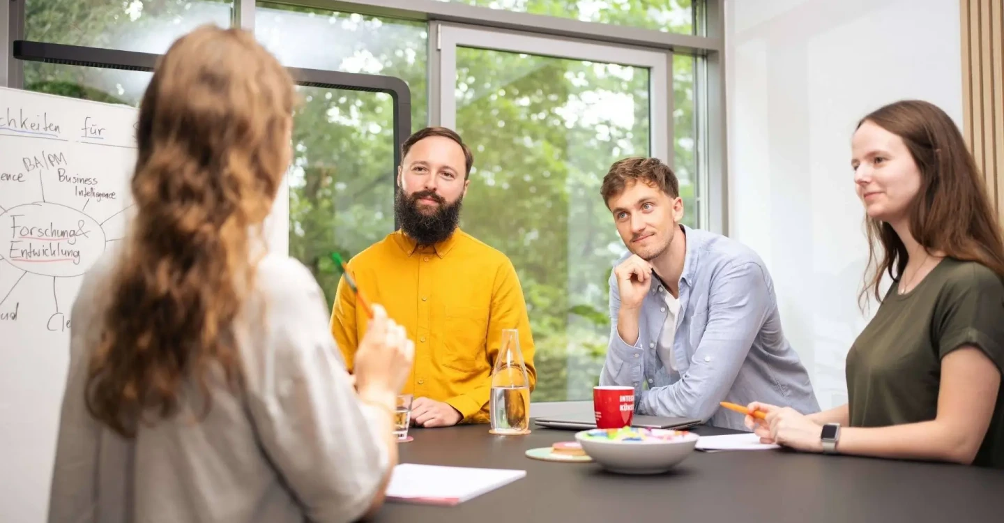 Drei Berater:innen der viadee stehen an einem Tisch und hören einer Werkstudentin zu.