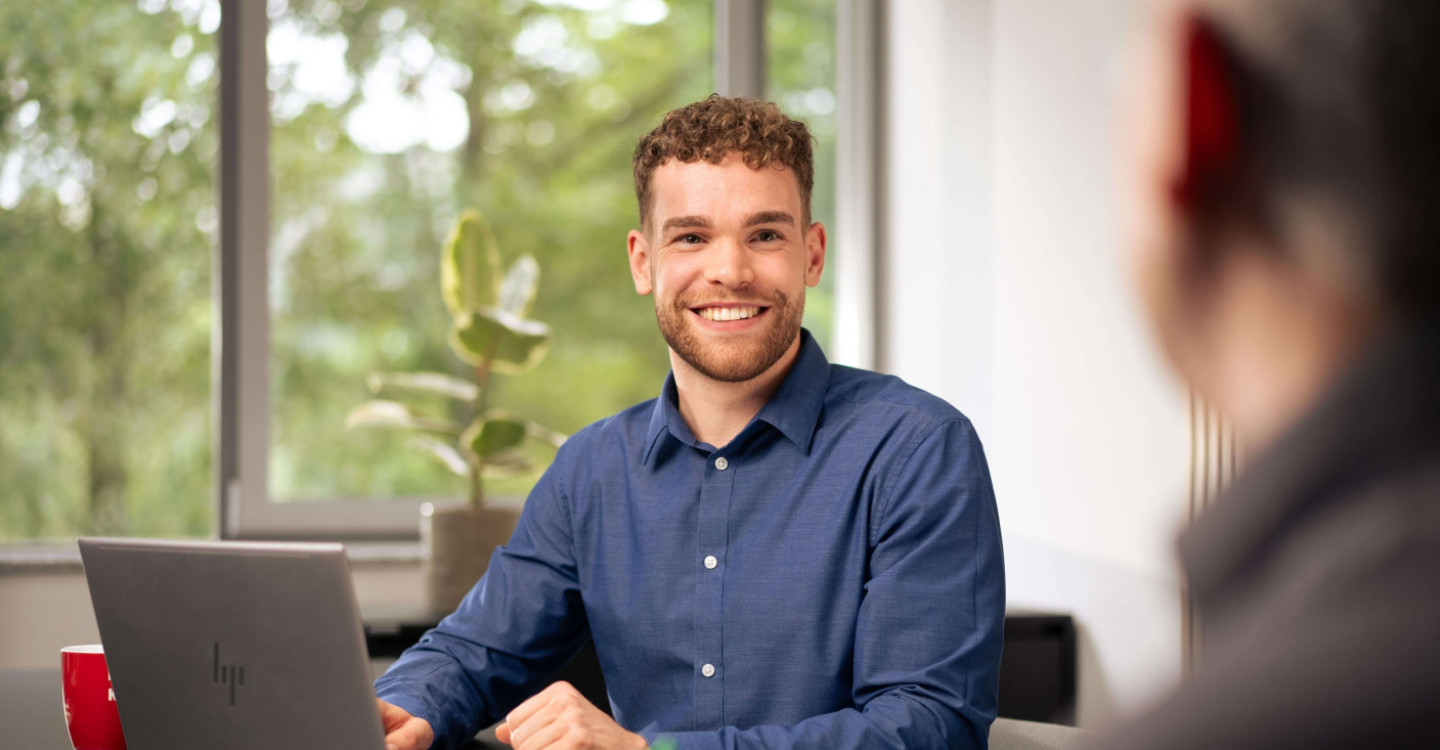 Ein Werkstudierender der viadee sitzt mit sinem Laptop an einem Schreibtisch und lächelt einen Kollegen an.