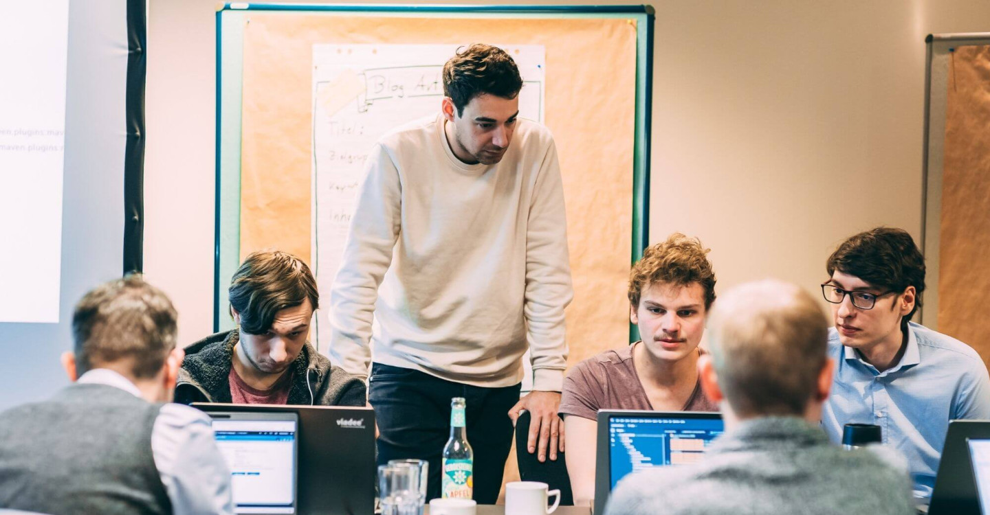 Fünf Mitarbeiter:innen der viadee sitzen um einen Tisch und arbeiten. Ein sechster Mitarbeiter steht bei ihnen .