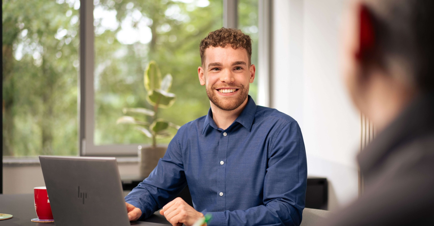 Ein Werkstudierender der viadee sitzt mit sinem Laptop an einem Schreibtisch und lächelt einen Kollegen an.