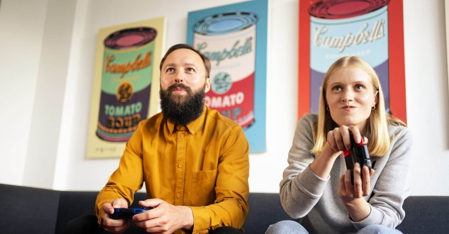 Eine Werkstudentin und ein Berater der viadee sitzen auf einer Couch. Sie spielen zusammen Videospiele.